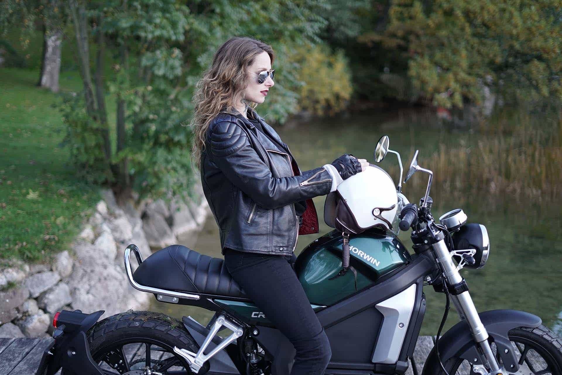 Lady straddling green Horwin motorcycle with white helmet by water