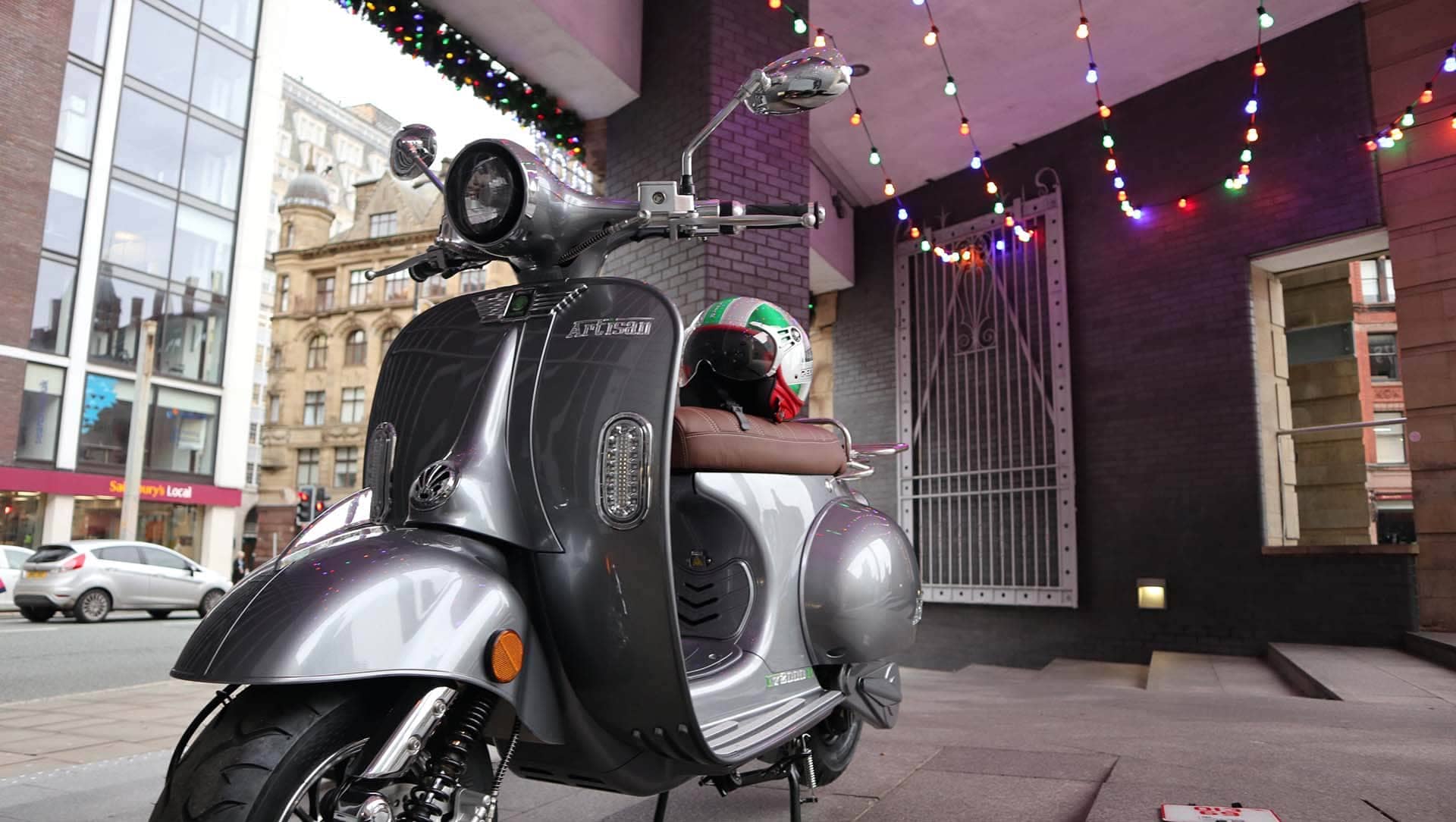 Grey Artisan scooter with helmet on the seat in a city with Christmas lights